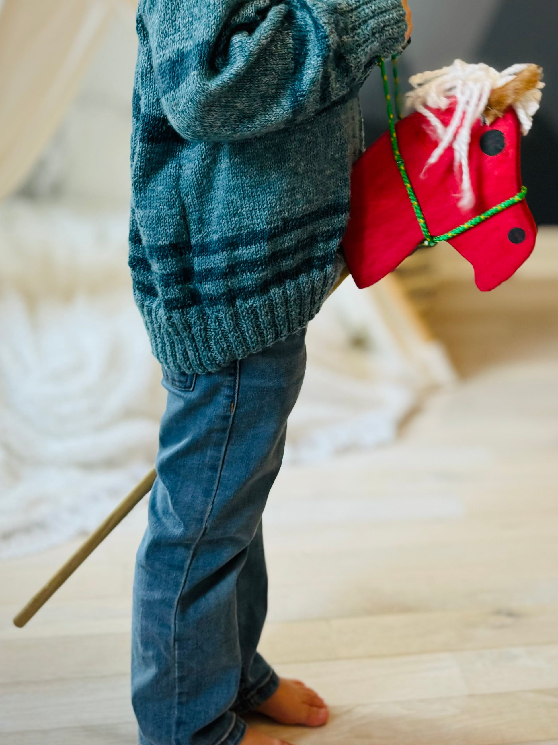 Child playing with red kids wooden hobby horses indoor wearing blue jeans and green sweater
