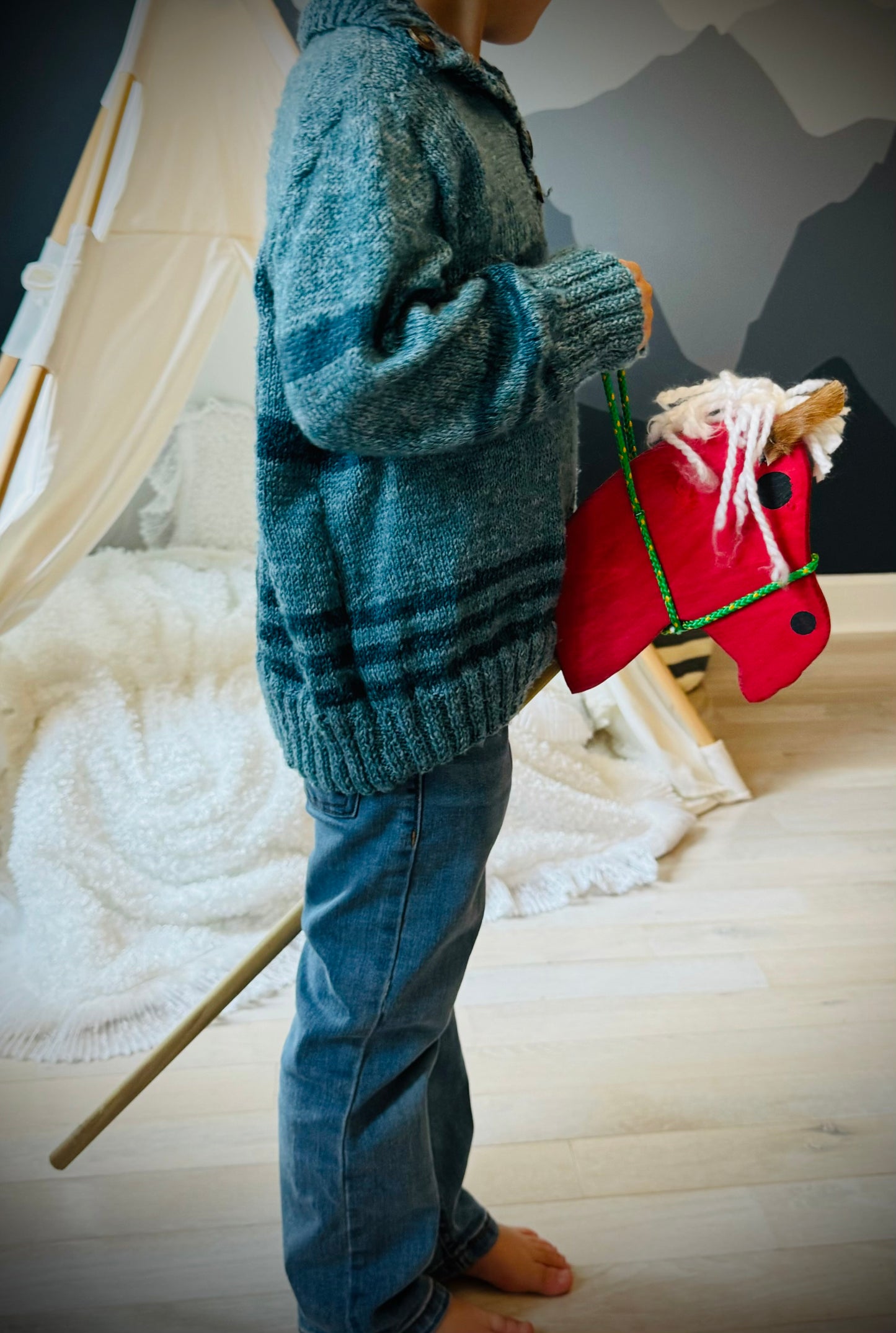 Child holding a red kids wooden hobby horse toy in a cozy indoor playroom