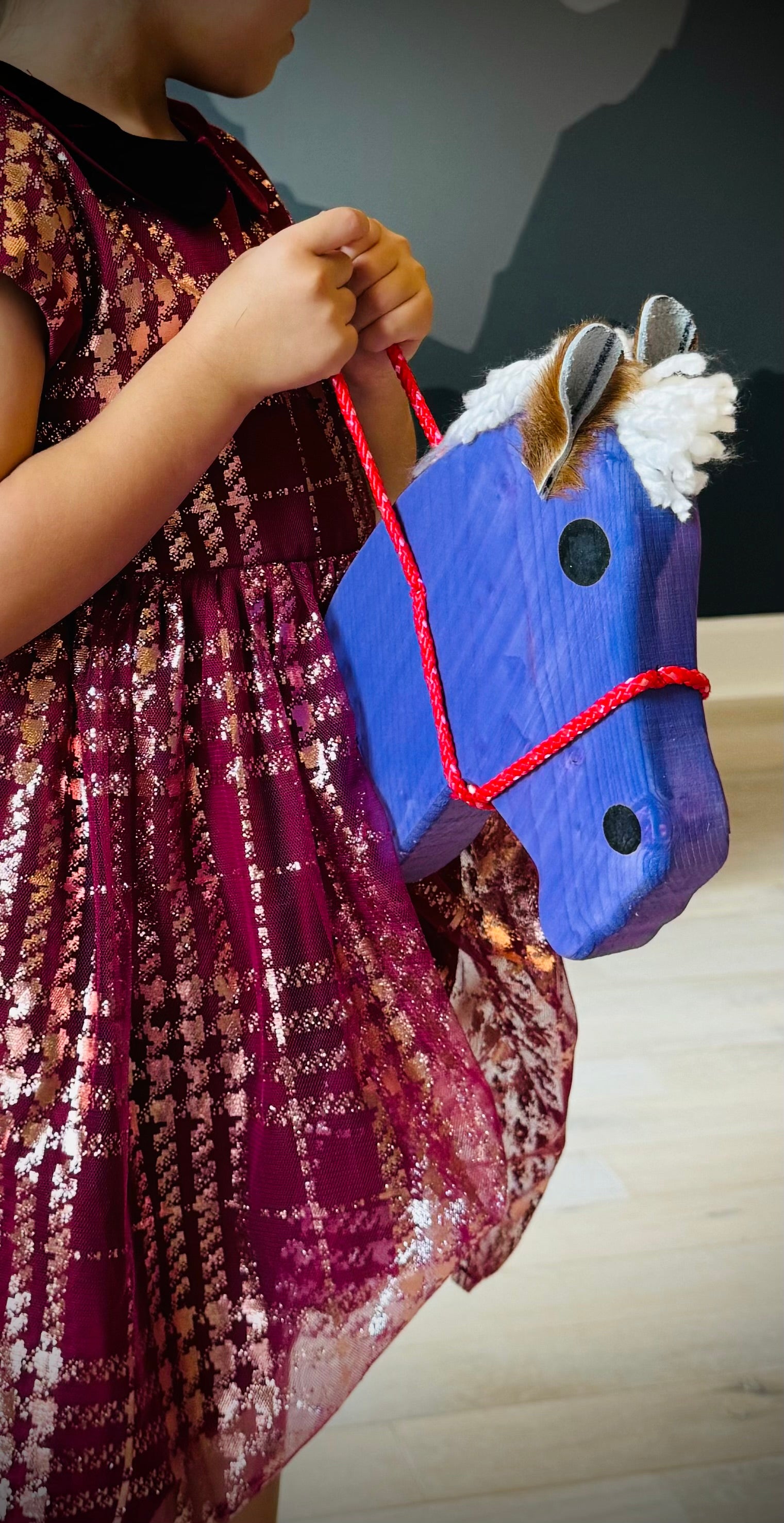 Child holding a blue kids wooden hobby horse with red reins and white mane indoors