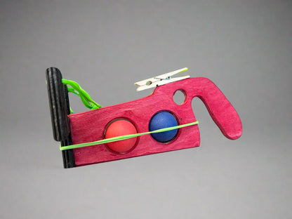 Kids wooden ping pong ball launcher toy in red with blue and orange balls and rubber band mechanism