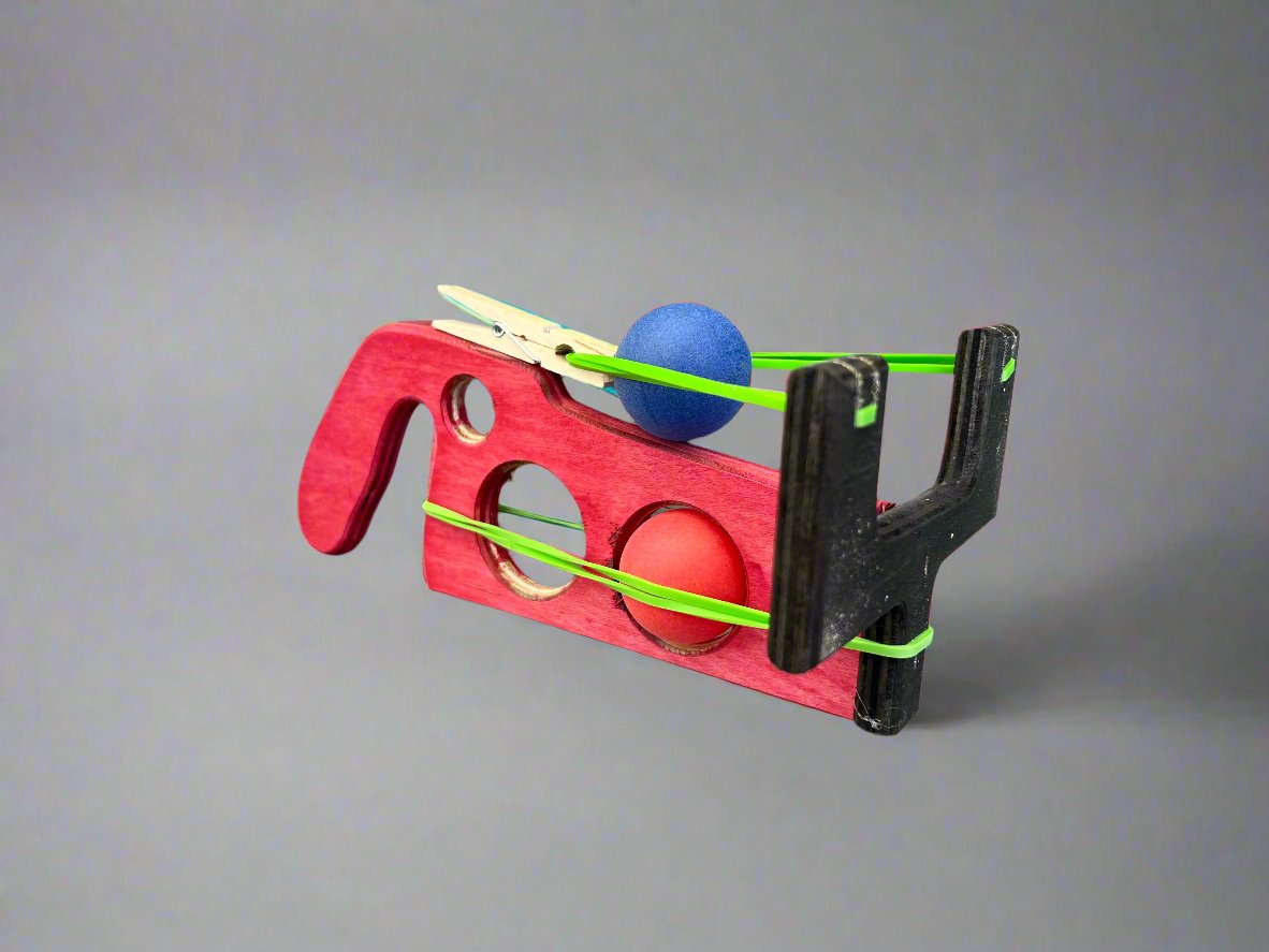 Kids wooden ping pong ball launcher in red and black with elastic bands and colorful balls on gray background