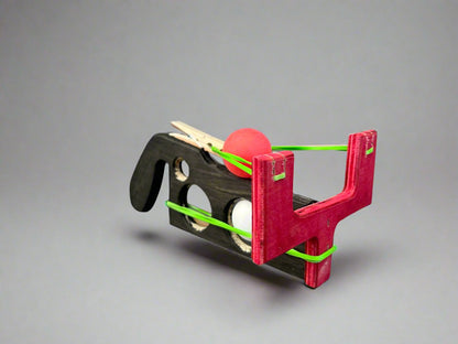 Kids wooden ping pong ball launcher with red and black wooden frame and green rubber bands