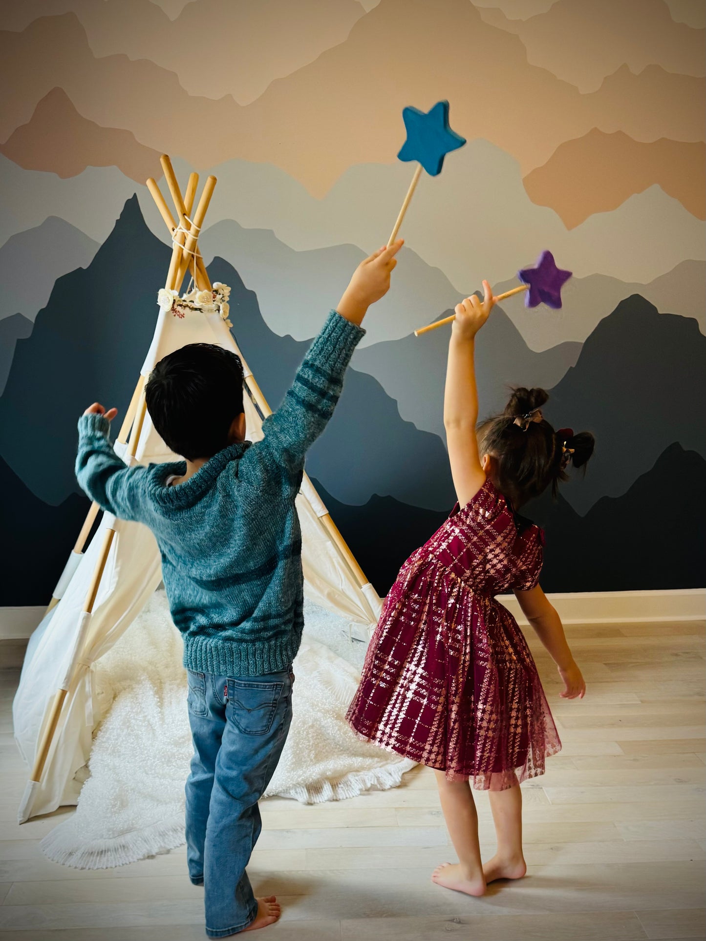 Two kids playing indoors with colorful wooden magic wand toy in front of a teepee and mountain wall mural