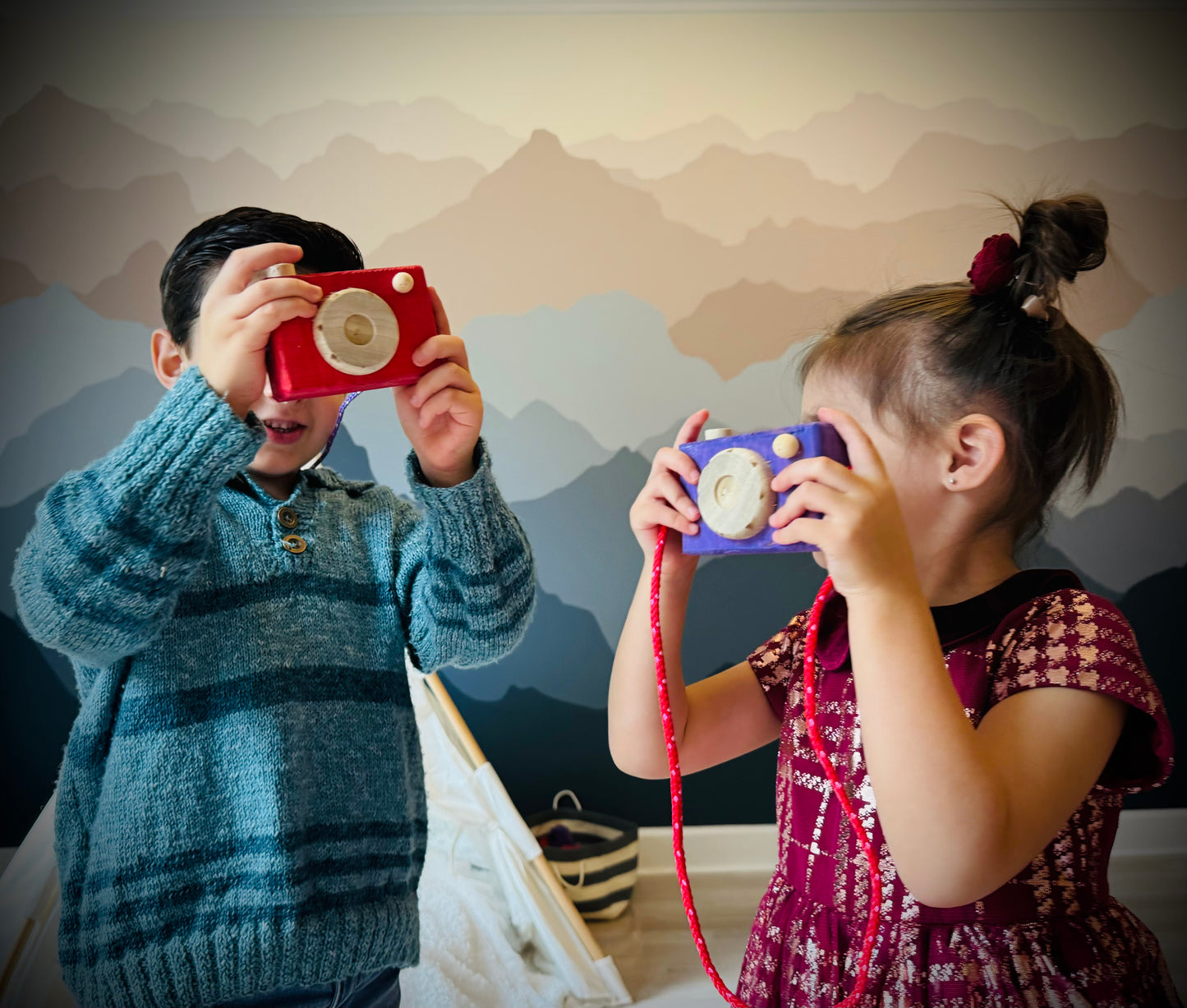 Children playing with colorful handmade kids wooden camera toys indoors with mountain mural background