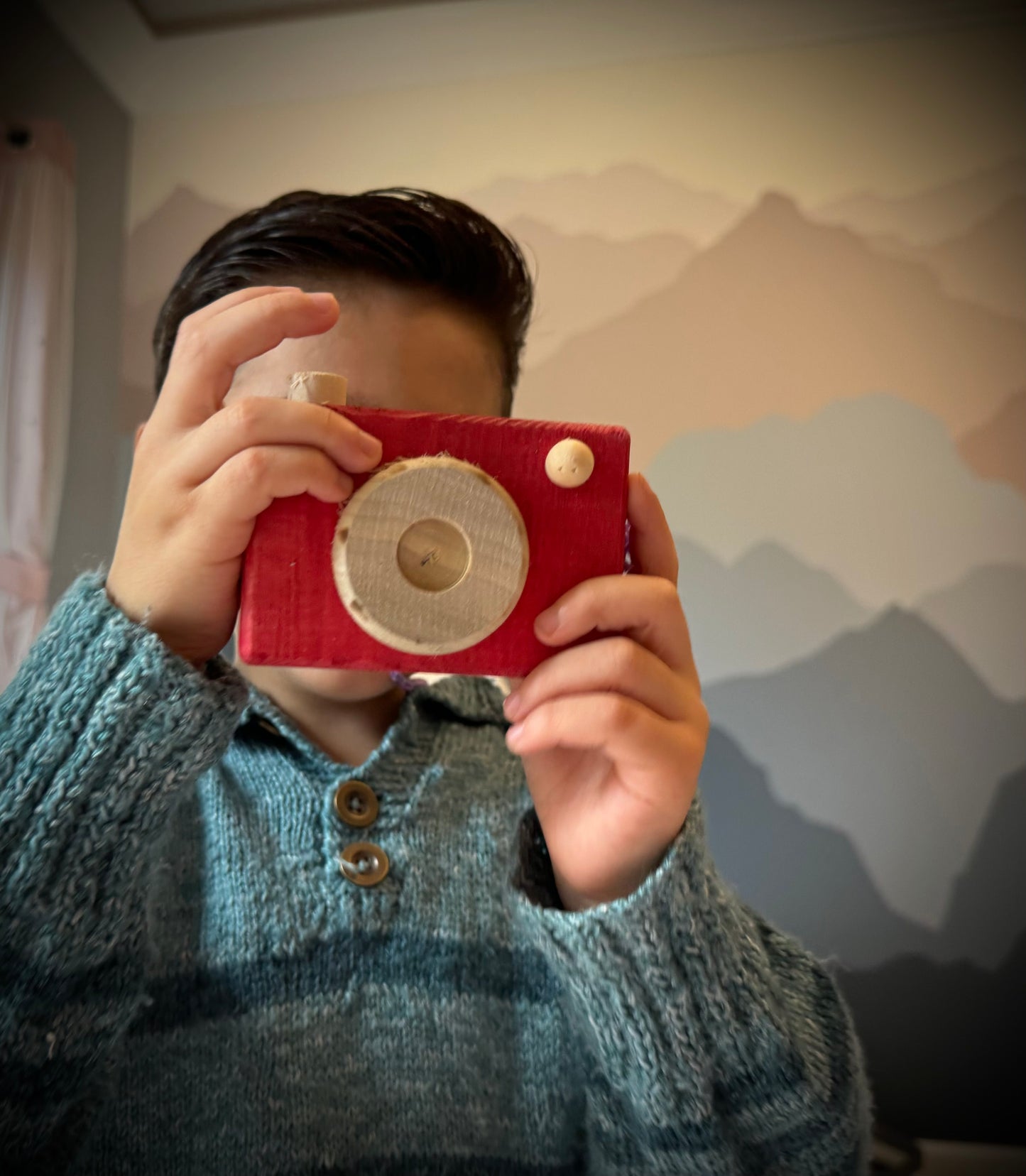 Child holding a handmade kids wooden camera in red, posing as if taking a photo indoors