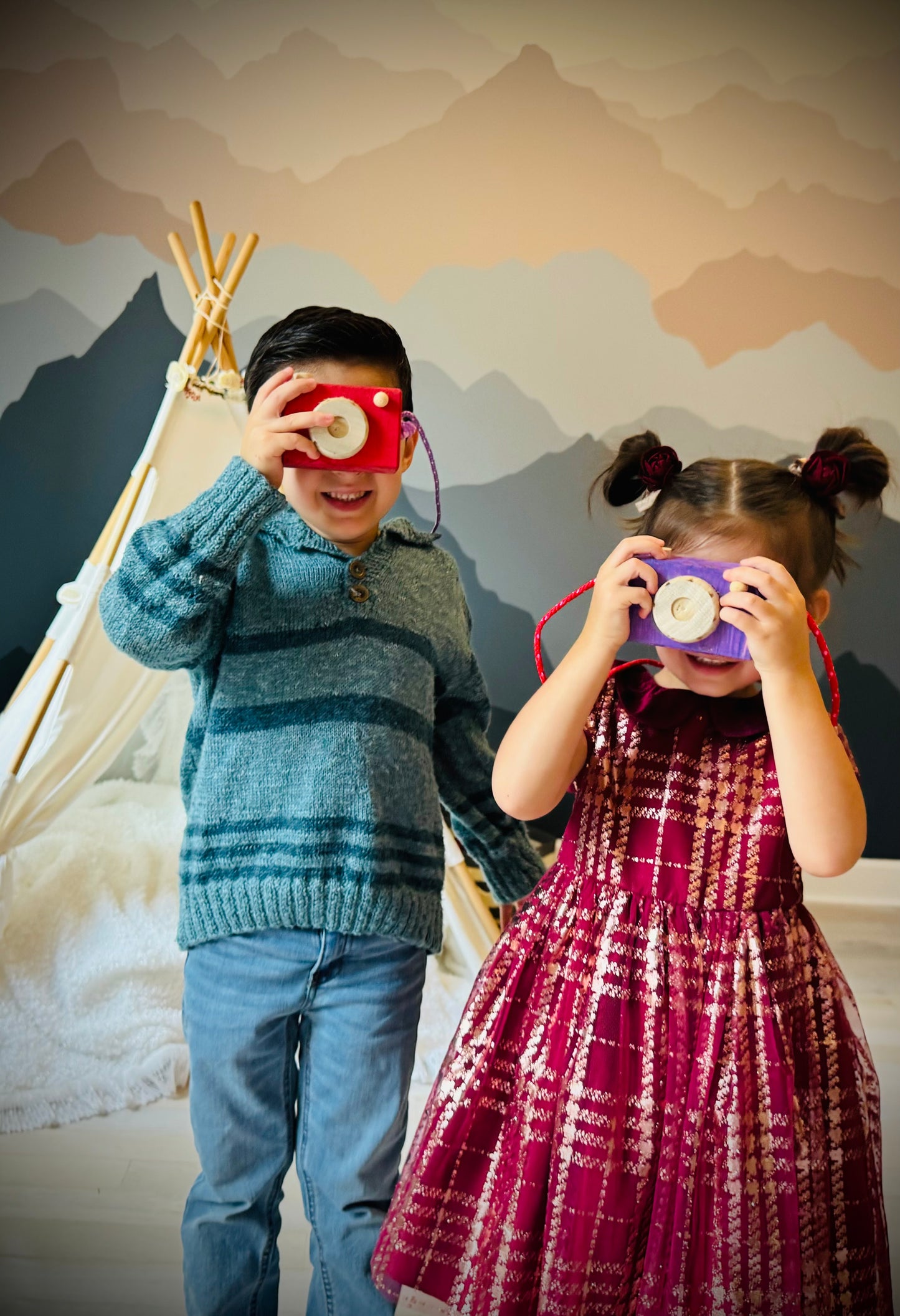 Two kids playing with a handmade kids wooden camera in a cozy indoor setting with mountain wall art