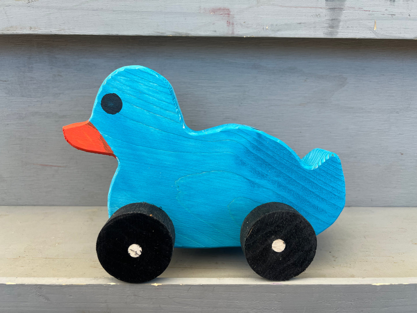Blue wooden pull duck toy with orange beak and black wheels on a gray surface
