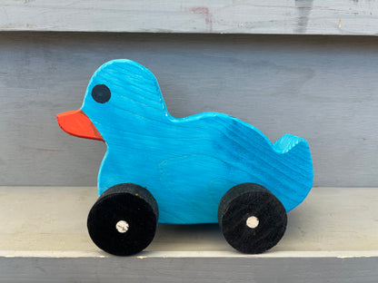 Blue wooden pull duck toy with orange beak and black wheels on a gray surface