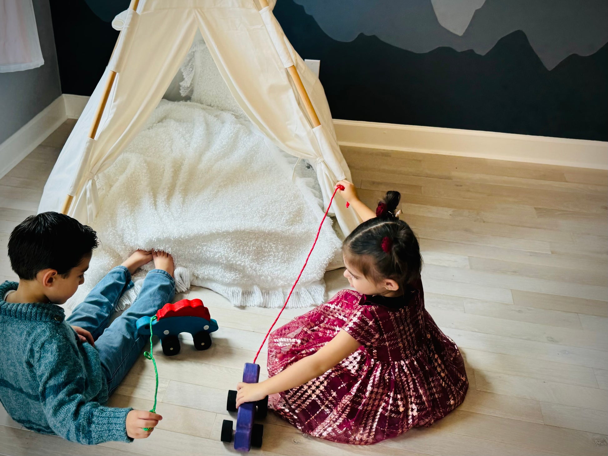 Two children playing with a wooden pull duck toy near a white play tent on wooden floor